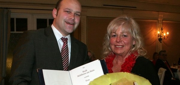 Bürgermeister Daniel Tybussek trägt einen Anzug. Er hält eine große Urkunde in der Hand und lächelt. Neben ihm steht eine Frau mit hellen Haaren und rotem Schal. Die Frau hält einen Blumenstrauß mit gelben Blumen. Beide wirken glücklich und stolz. Im Hintergrund sieht man einen festlich geschmückten Raum mit warmem Licht.