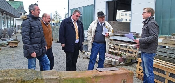 Das Bild zeigt fünf Männer, die sich draußen auf einem gepflasterten Platz versammelt haben. In der Mitte liegt ein großer, länglicher Steinblock auf dem Boden. Der Stein hat eine rötlich-grüne Farbe und eine raue Oberfläche. Die Männer stehen um den Stein herum und betrachten ihn aufmerksam. Der Mann rechts hält Unterlagen in den Händen. Die anderen Männer tragen warme Kleidung wie Jacken und Mäntel. Im Hintergrund sind Paletten mit weiteren Steinen zu sehen. Auch ein Gebäude mit großen Fenstern ist zu sehen. Die Stimmung wirkt konzentriert und interessiert.
