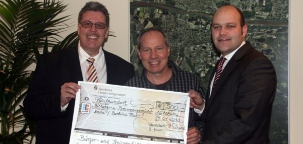 Drei Männer stehen gemeinsam in einem Raum. Sie halten einen großen, symbolischen Scheck in den Händen. Der Scheck zeigt einen Betrag von 500 Euro. Die Männer lächeln freundlich in die Kamera. Links steht ein Mann im Anzug mit Krawatte. In der Mitte ein Mann im karierten Hemd. Rechts steht Bürgermeister Daniel Tybussek im Anzug. Im Hintergrund ist ein großes Bild von einer Karte zu sehen, die Mühlheim am Main von oben zeigt. Die Atmosphäre wirkt positiv und festlich.