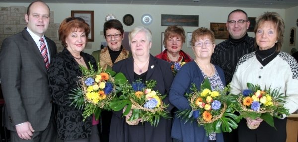 Das Bild zeigt acht Personen, die gemeinsam in einem hellen Raum stehen. Fünf Frauen in der Mitte halten bunte Blumensträuße in den Händen. Die Sträuße sind mit gelben, blauen und orangefarbenen Blumen geschmückt. Links und rechts neben den Frauen stehen zwei Männer. Unter ihnen Bürgermeister Daniel Tybussek. Im Hintergrund sind helle Wände, Tische, Stühle und Dekorationen an der Wand zu sehen. Die Stimmung wirkt feierlich und fröhlich.