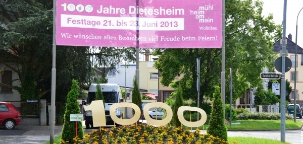 Auf dem Bild sieht man eine grüne Wiese mit vielen bunten Blumen. In der Mitte stehen große gelbe Zahlen: 1000. Über der Wiese hängt ein großes, pinkes Schild. Auf dem Schild steht: "1000 Jahre Dietesheim". Darunter steht: "!Festtage 21. bis 23. Juni 2013". Es wird allen Besuchern viel Freude beim Feiern gewünscht. Im Hintergrund sind Bäume, Häuser und ein weißer Transporter zu sehen.