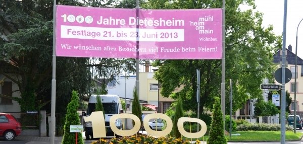 Auf dem Bild sieht man eine grüne Wiese mit vielen bunten Blumen. In der Mitte stehen große gelbe Zahlen: 1000. Über der Wiese hängt ein großes, pinkes Schild. Auf dem Schild steht: "1000 Jahre Dietesheim". Darunter steht: "!Festtage 21. bis 23. Juni 2013". Es wird allen Besuchern viel Freude beim Feiern gewünscht. Im Hintergrund sind Bäume, Häuser und ein weißer Transporter zu sehen.