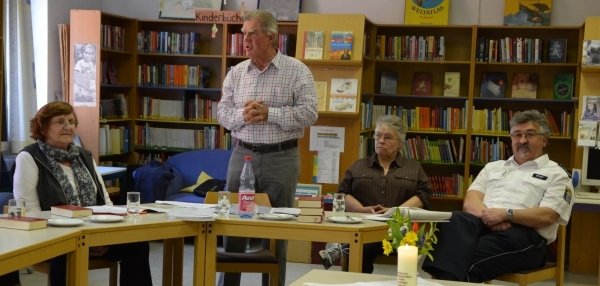 Auf dem Bild sind vier Personen zu sehen, die an einem Tisch sitzen. Eine Person steht und spricht zu den anderen. Im Hintergrund sind viele Bücher in Regalen. Auf dem Tisch stehen Getränke, Bücher und in der Mitte eine brennende Kerze.