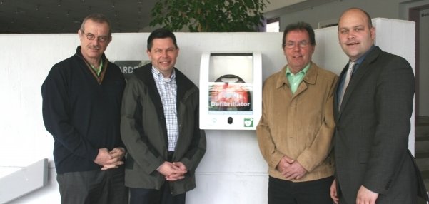 Vier Männer stehen vor einer weißen Wand. An der Wand hängt ein Defibrillator. Ein Defibrillator ist ein Gerät, das bei Herznotfällen helfen kann. Die Männer lächeln in die Kamera. Im Hintergrund ist ein grüner Baum zu sehen. Der Raum wirkt hell und freundlich.