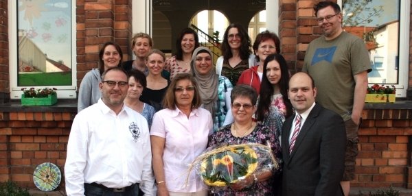 Viele Menschen stehen vor einem Gebäude aus roten Ziegelsteinen. Eine Frau in der Mitte hält einen Blumenstrauß in den Händen. Alle lächeln freundlich in die Kamera. Im Hintergrund sind große Fenster und eine geöffnete Tür zu sehen. Links und rechts stehen grüne Pflanzen in Töpfen.