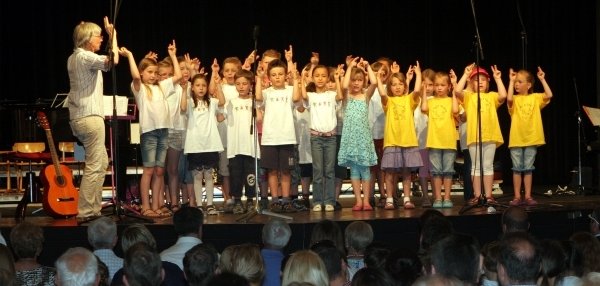 Auf dem Bild steht eine große Gruppe von Kindern auf  einer Bühne. Die Kinder tragen weiße und gelbe T-Shirts und halten die Arme nach oben. Eine Frau vorne links leitet die Kinder an. Vor der Bühne sitzen viele Erwachsene und schauen zu. Rechts steht eine Gitarre auf der Bühne.