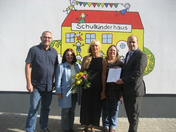 Fünf Personen stehen zusammen vor einer bunten Wand mit einem gemalten Schulhaus. Eine Frau hält einen Blumenstrauß in der Hand. Eine andere Person hält ein Dokument- Alle schauen freundlich in die Kamera.