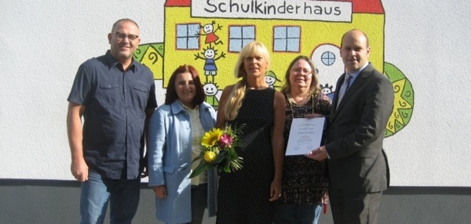Fünf Personen stehen zusammen vor einer bunten Wand mit einem gemalten Schulhaus. Eine Frau hält einen Blumenstrauß in der Hand. Eine andere Person hält ein Dokument- Alle schauen freundlich in die Kamera.