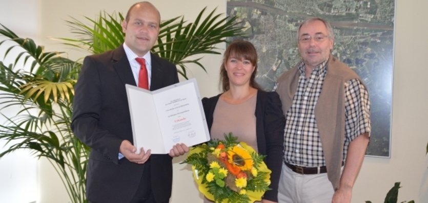 Drei Personen stehen gemeinsam vor einer hellen Wand mit grünen Pflanzen. Links im Bild steht Bürgermeister Daniel Tybussek. Er hält eine große Urkunde in den Händen. Neben ihm steht eine Frau mit schulterlangem Haar. Sie hält einen bunten Blumenstrauß mit gelben und orangefarbenen Blumen. Rechts daneben steht ein weiterer Mann. Im Hintergrund ist ein großes Bild an der Wand zu sehen. Es zeigt die Stadtansicht von Mühlheim am Main. Die Stimmung wirkt freundlich und feierlich.