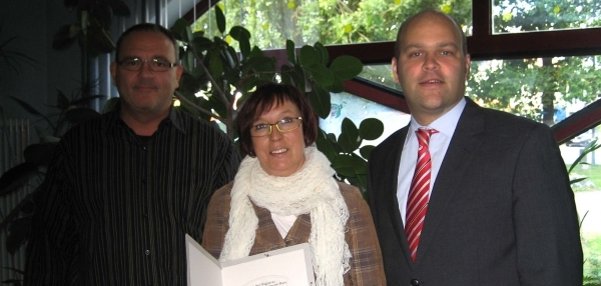 Drei Personen stehen nebeneinander vor einem großen Fenster mit grünen Pflanzen im Hintergrund. Die Frau in der Mitte hält eine Urkunde in der einen Hand. In der anderen Hand hält sie einen bunten Blumenstrauß. Sie lächelt freundlich in die Kamera. Links neben ihr steht  ein Mann mit Brille. Recht neben ihr steht Bürgermeister Daniel Tybussek. Die Atmosphäre wirkt fröhlich und feierlich.