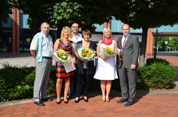 Sechs Personen stehen nebeneinander auf einem gepflasterten Platz im Freien. Hinter ihnen sind Bäume und ein Gebäude mit großen Fenstern zu sehen. Drei Frauen in der Mitte halten bunte Blumensträuße in den Händen. Sie lächeln freundlich. Die Männer links und rechts neben ihnen schauen ebenfalls in die Kamera. Einer von ihnen ist Bürgermeister Daniel Tybussek. Die Stimmung ist fröhlich.