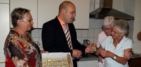 Auf dem Bild stehen vier Personen in einer Küche. Der Mann in der Mitte ist Bürgermeister Daniel Tybussek. er hält eine Schachtel in der Hand. Die Frau links hält eine Box mit weiteren Präsenten in der Hand. Alle schauen interessiert auf die Präsente und lächeln. Auf dem Küchentisch stehen Gläser mit Getränke. Im Hintergrund ist eine Kücheneinrichtung zu sehen.