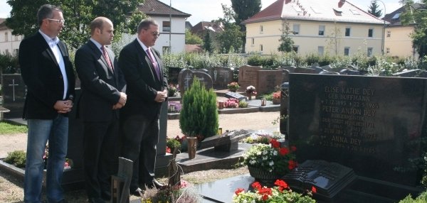 Drei Männer stehen an einem Grab auf einem Friedhof. Darunter Bürgermeister Daniel Tybussek. Sie schauen ernst und respektvoll auf den großen Grabstein. Vor dem Grabstein stehen viele bunte Blumen und Pflanzen. Im Hintergrund sind weitere Gräber sowie Häuser mit roten Dächern zu sehen. Die Männer haben die Hände vor dem Körper gefaltet. Die Atmosphäre ist ruhig und würdevoll.