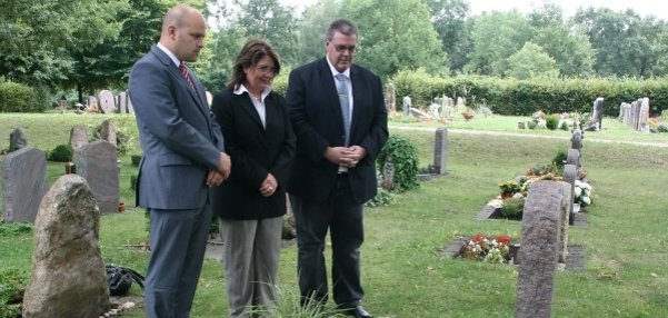 Drei Personen stehen an einem Grab auf einem grünen Friedhof. Vor dem Grabstein liegen bunte Blumen und ein Krank mit einer Schleife. Die drei Personen, darunter Bürgermeister Daniel Tybussek, schauen respektvoll auf das Grab. Sie haben die Hände vor dem Körper gefaltet. Im Hintergrund sind weitere Grabsteine und viele Bäume zu sehen. Die Stimmung ist still und ehrwürdig.