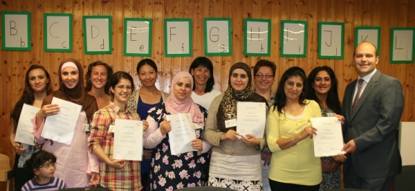 Viele Frauen stehen zusammen und halten Zertifikate in den Händen. Alle schauen freundlich in die Kamera. Im Hintergrund hängen grüne Buchstaben an der Holzwand. Ein Mann im Anzug, Bürgermeister Daniel Tybussek, steht am rechten Rand der Gruppe. Die Frauen wirken stolz und glücklich.