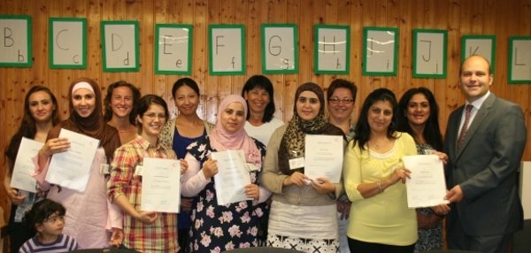 Viele Frauen stehen zusammen und halten Zertifikate in den Händen. Alle schauen freundlich in die Kamera. Im Hintergrund hängen grüne Buchstaben an der Holzwand. Ein Mann im Anzug, Bürgermeister Daniel Tybussek, steht am rechten Rand der Gruppe. Die Frauen wirken stolz und glücklich.