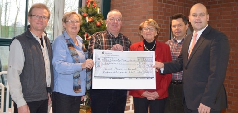 Sechs Personen stehen zusammen und halten einen großen symbolischen Scheck. Alle lächeln freundlich. Im Hintergrund ist ein weihnachtlich geschmückter Tannenbaum mit roten und goldenen Kugeln zu sehen. Im Hintergrund sind rote Backsteinwände und eine große Fensterfront zu sehen.