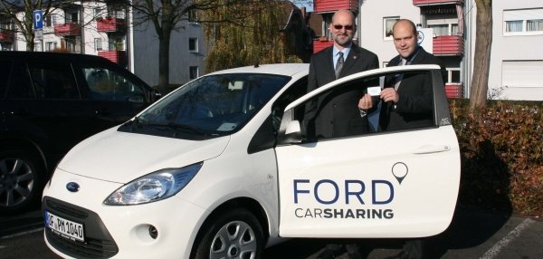Zwei Männer stehen neben einem weißen Auto mit der Aufschrift: "FORD CARSHARING". Einer der Männer ist Bürgermeister Daniel Tybussek. Beide Männer lächeln und halten einen Autoschlüssel in den Händen. Im Hintergrund sind Wohnhäuser mit roten Balkonen und herbstlich gefärbte Bäume zu sehen. Die Sonne scheint und wirft Schatten.