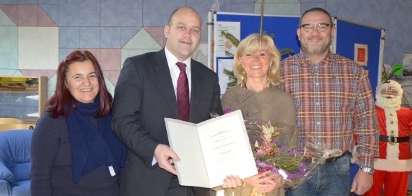 Auf dem Bild stehen vier Personen nebeneinander und lächeln in die Kamera. Der Mann im Anzug ist Bürgermeister Daniel Tybussek. Er hält eine Urkunde in der Hand. Die Frau neben ihm trägt einen Blumenstrauß. Die anderen beiden Personen stehen links und rechts daneben. Im Hintergrund ist eine bunte Wand und ein dekorierter Weihnachtsmann zu sehen.