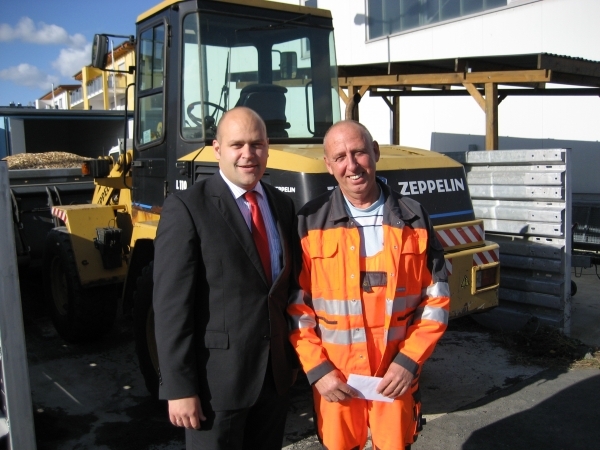 Auf dem Bild stehen zwei Männer draußen nebeneinander und lächeln in die Kamera. Der Mann links trägt einen dunklen Anzug mit roter Krawatte. Es ist Bürgermeister Daniel Tybussek. Der Mann rechts hat eine leuchtend orange Arbeitskleidung an. Er hält einen Umschlag in der Hand. Im Hintergrund steht eine große gelbe Baumaschine. Die Sonne scheint.