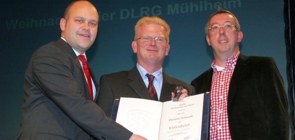 Drei Männer stehen nebeneinander und lächeln in die Kamera. Der Mann links trägt einen dunklen Anzug mit roter Krawatte. Es ist Bürgermeister Daniel Tybussek. Er hält zusammen mit dem Mann in der Mitte eine Urkunde. Der Mann rechts trägt ein rotes kariertes Hemd und eine schwarze Jacke. Im Hintergrund ist eine blaue Wand mit Schriftzug zu sehen.