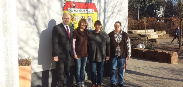 Vier Personen stehen nebeneinander vor einer weißen Wand. Auf der Wand ist ein buntes Bild mit der Aufschrift "Schulkinderhaus" gemalt. Links steht ein Mann im Anzug und roter Krawatte. Es ist Bürgermeister Daniel Tybussek. Daneben stehen drei Frauen in Alltagskleidung. Alle lächeln freundlich in die Kamera. Im Hintergrund sind Bäume, Pflanzen und ein kleiner gepflasterter Platz zu sehen, 