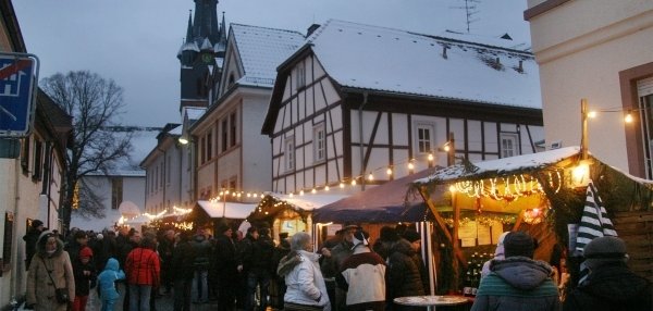 Das Foto zeigt eine winterliche Szene in einer schmalen Straße mit vielen Menschen. Die Straße ist mit kleinen Holzhütten gesäumt, die festlich mit Lichtern und Dekorationen geschmückt sind. Viele Menschen stehen an den Ständen und schauen sich um. Im Hintergrund steht ein großes Fachwerkhaus mit einem spitzen Kirchturm. Auf den Dächern liegt Schnee. Es ist bereits dämmrig. 