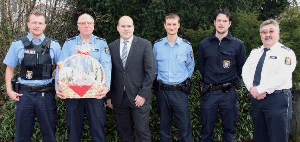 Auf dem Foto stehen sechs Männer nebeneinander vor grünen Büschen. Vier der Männer tragen Polizeiuniformen in hellblau und dunkelblau. In der Mitte steht Bürgermeister Daniel Tybussek. Er trägt einen grauen Anzug mit Krawatte. Der Polizist links hält einen großen Präsentkorb mit einer roten Schleife. Alle lächeln freundlich in die Kamera. Im Hintergrund sind Bäume und Büsche zu sehen.