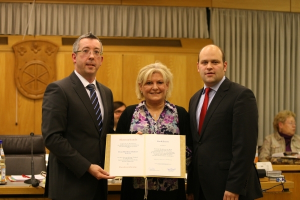 Das Foto zeigt drei Personen in einem Raum mit Holzvertäfelung. In der Mitte steht eine Frau mit blonden Haaren. Sie hält ein offenes Dokument in den Händen. Links und rechts von ihr stehen zwei Männer in Anzügen. Einer von ihnen ist Bürgermeister Daniel Tybussek. Alle drei blicken freundlich in die Kamera. Im Hintergrund sind weitere sitzende Personen und ein Wappen an der Wand zu sehen.