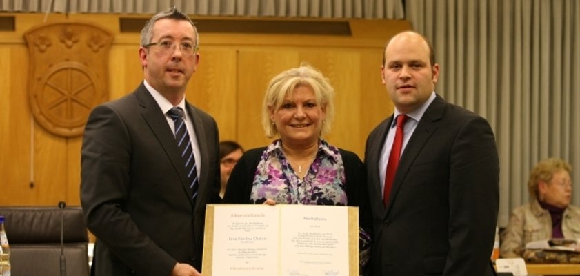 Das Foto zeigt drei Personen in einem Raum mit Holzvertäfelung. In der Mitte steht eine Frau mit blonden Haaren. Sie hält ein offenes Dokument in den Händen. Links und rechts von ihr stehen zwei Männer in Anzügen. Einer von ihnen ist Bürgermeister Daniel Tybussek. Alle drei blicken freundlich in die Kamera. Im Hintergrund sind weitere sitzende Personen und ein Wappen an der Wand zu sehen.