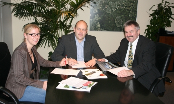 Drei Personen sitzen an einem dunklen Tisch. Links ist eine blonde Frau mit Brille. In der Mitte sitzt Bürgermeister Daniel Tybussek. Er trägt einen Anzug und schreibt gerade etwas. Rechts sitzt ein weiterer Mann im Anzug. Er hält ein Dokument in der Hand. Auf dem Tisch liegen verschiedene Unterlagen und Broschüren. Im Hintergrund sind große Pflanzen und ein Bild von einer Stadtansicht an der Wand.