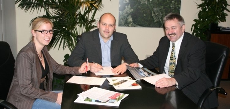 Drei Personen sitzen an einem dunklen Tisch. Links ist eine blonde Frau mit Brille. In der Mitte sitzt Bürgermeister Daniel Tybussek. Er trägt einen Anzug und schreibt gerade etwas. Rechts sitzt ein weiterer Mann im Anzug. Er hält ein Dokument in der Hand. Auf dem Tisch liegen verschiedene Unterlagen und Broschüren. Im Hintergrund sind große Pflanzen und ein Bild von einer Stadtansicht an der Wand.