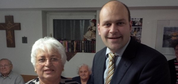 Auf dem Bild sind zwei Personen zu sehen. Links steht eine ältere Frau mit weißem Haar und einer schwarz-weiß gestreiften Jacke- Sie lächelt freundlich. Rechts neben ihr steht Bürgermeister Daniel Tybussek. Er trägt einen Anzug und hält eine Urkunde. Im Hintergrund sitzen weitere Menschen an einem Tisch. An der Wand hängt ein großes Holzkreuz und Bilder.