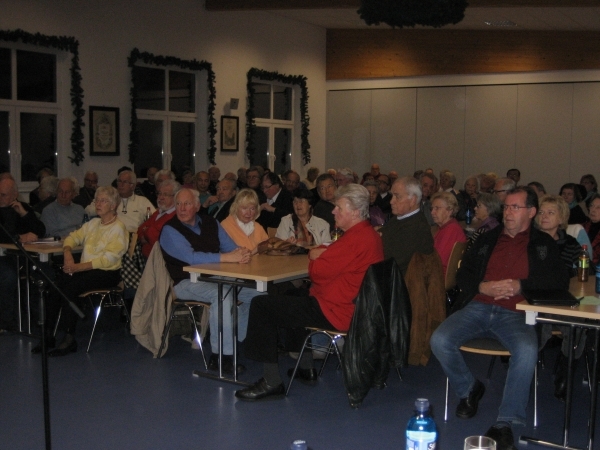 Das Bild zeigt einen großen Raum, der voller Menschen ist. Viele der Anwesenden sind ältere Personen. Sie sitzen an Tischen. An den Wänden hängen Bilder und die Fenster sind mit grüner Dekoration geschmückt. Auf den Tischen stehen Wasserflaschen und Gläser.