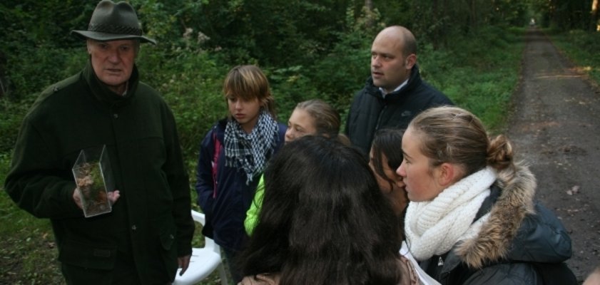 Das Bild zeigt eine Gruppe von Menschen in einem Wald. Ein älterer Mann mit Hut und dunkler Jacke hält ein durchsichtiges Behältnis in der Hand. Um ihn herum stehen mehrere Kinder und Erwachsene. Im Hintergrund sind viele grüne Bäume und ein Waldweg zu sehen. Die Menschen tragen warme Jacken.