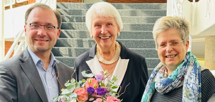 Das Bild zeigt drei Personen, die gemeinsam lächeln. Links steht Bürgermeister Dr. Alexander Krey. In der Mitte steht eine Frau mit kurzen grauen Haaren, die einen Blumenstrauß hält. Rechts steht Gudrun Monat mit einer Urkunde. Sie ist jetzt ehrenamtliche Jumelage-Beauftragte der Stadt Mühlheim. Das bedeutet, sie kümmert sich um die Städtepartnerschaft mit Saint-Priest. Im Hintergrund sieht man eine graue Treppe in einem Gebäude.