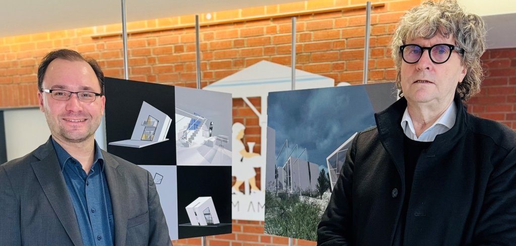 Das Bild zeigt zwei Männer in einem Gebäude mit roten Backsteinwänden. Links steht Bürgermeister Dr. Alexander Krey in einem dunklen Anzug und lächelt. Rechts steht Architekt Gerald Marx mit lockigen Haaren und schwarzer Kleidung. Hinter ihnen sind Tafeln mit Architekturentwürfen zu sehen. In der Mitte steht ein kleines Modell eines Gebäudes auf einem Podest.