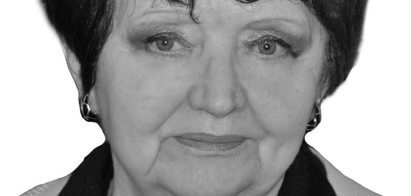 Das Bild zeigt eine ältere Frau mit kurzen, dunklen Haaren. Sie trägt ein schwarzes Oberteil mit und eine weiße Strickjacke. Sie trägt Ohrringe. Ihr Gesichtsausdruck ist ruhig und ernst. Das Foto ist in Schwarz-Weiß und zeigt sie vor einem neutralen Hintergrund.