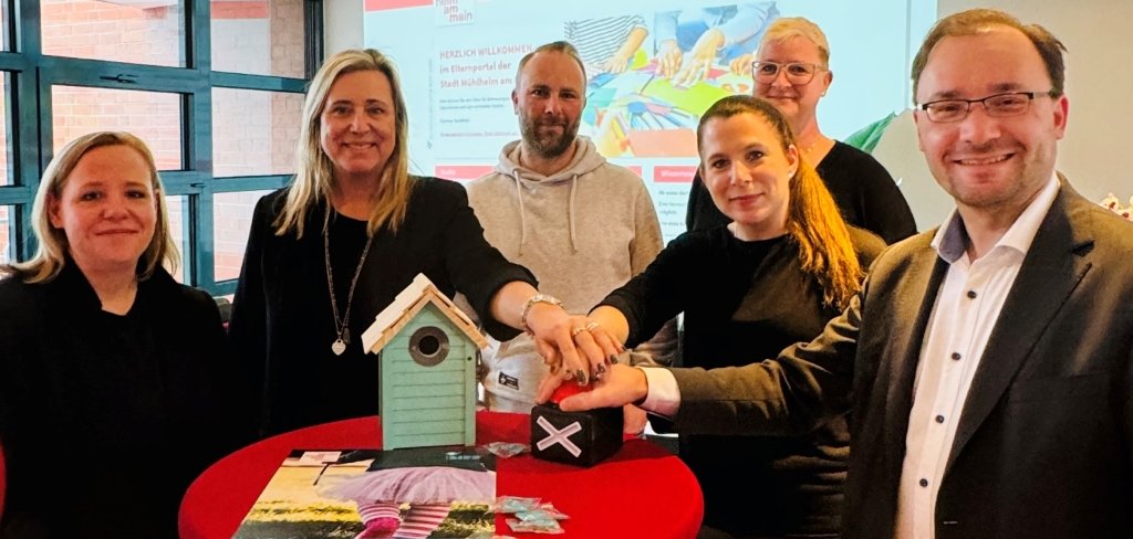 Das Bild zeigt sechs Personen, die gemeinsam auf einen Knopf drücken. Der Bürgermeister steht rechts und lächelt. Auf dem Tisch vor ihnen steht ein kleines, buntes Vogelhäuschen. Im Hintergrund ist eine große Leinwand mit einer Internetseite zu sehen.