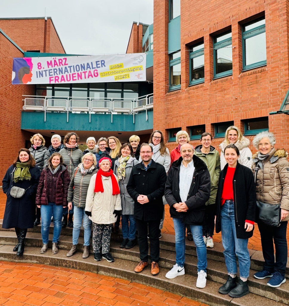 Gruppenbild vor dem neuen Banner anlässlich des internationalen Frauentages Das Bild zeigt eine Gruppe von Menschen vor einem roten Backsteingebäude. Über ihnen hängt ein großes Banner mit der Aufschrift "8. März - Internationaler Frauentag". In der Mitte steht der Bürgermeister Dr. Alexander Krey, umgeben von vielen Frauen. Alle tragen warme Kleidung. Sie lächeln und schauen in die Kamera.