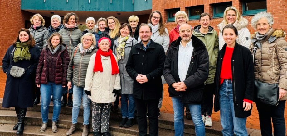 Das Bild zeigt eine Gruppe von Menschen vor einem roten Backsteingebäude. Über ihnen hängt ein großes Banner mit der Aufschrift "8. März - Internationaler Frauentag". In der Mitte steht der Bürgermeister Dr. Alexander Krey, umgeben von vielen Frauen. Alle tragen warme Kleidung. Sie lächeln und schauen in die Kamera.