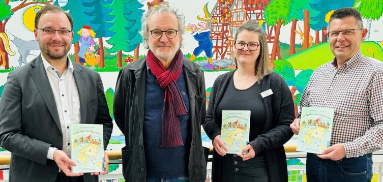 Vier Personen stehen in einem Gebäude vor einer bunten Wandmalerei mit Märchenmotiven. Sie halten Bücher mit dem Titel "Die Abenteuer des Simplicissimus" in der Hand. Links steht Bürgermeister Dr. Alexander Krey im Anzug. In der Mitte steht Klaus Puth, der Illustrator des Buches. Rechts neben ihm steht eine Frau mit Brille. Sie ist die Leiterin der Stadtbücherei. Ganz rechts hält ein Mann mit kariertem Hemd ebenfalls ein Buch in der Hand. Er ist der Fachbereichsleiter. Alle lächeln in die Kamera.