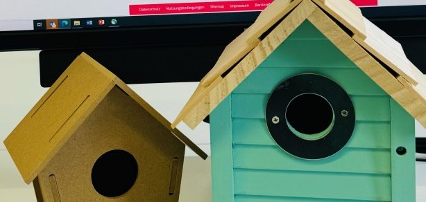 Das Bild zeigt einen Computerbildschirm mit der Website des digitalen Kita-Anmeldeportals der Stadt Mühlheim. Auf dem Tisch davor stehen zwei Vogelhäuschen. Eines in braun, eines in türkis. Das braune Häuschen hat die Aufschrift "Little Bird". Außerdem liegen kleine verpackte Bonbons mit der Aufschrift "House" auf dem Tisch.