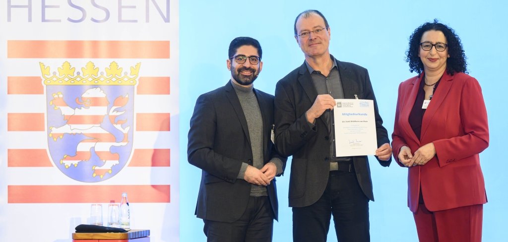 Das Bild zeigt drei Personen auf einer Bühne. In der Mitte hält Erster Stadtrat Robert Ahrnt eine Urkunde in der Hand und lächelt. Links neben ihm steht ein Mann mit Brille und Bart, rechts eine Frau in einem roten Anzug. Hinter ihnen ist eine große Fahne mit dem Wappen von Hessen zu sehen. Die Stadt Mühlheim ist jetzt Mitglied in der Arbeitsgemeinschaft Nahmobilität Hessen.