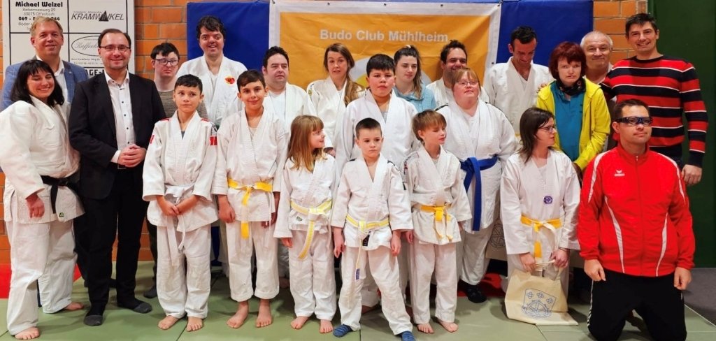 Das Bild zeigt eine große Gruppe von Menschen in einer Sporthalle. Viele tragen weiße Judoanzüge mit verschiedenen Gürtelfarben. Im Hintergrund hängt ein Banner mit der Aufschrift "50 Jahre Budo Sport in Mühlheim 1972 - 2022". Bürgermeister Dr. Alexander Krey steht links in dunkler Kleidung und lächelt. Alle schauen in die Kamera.