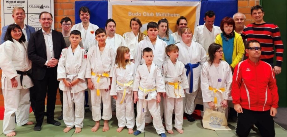 Das Bild zeigt eine große Gruppe von Menschen in einer Sporthalle. Viele tragen weiße Judoanzüge mit verschiedenen Gürtelfarben. Im Hintergrund hängt ein Banner mit der Aufschrift "50 Jahre Budo Sport in Mühlheim 1972 - 2022". Bürgermeister Dr. Alexander Krey steht links in dunkler Kleidung und lächelt. Alle schauen in die Kamera.