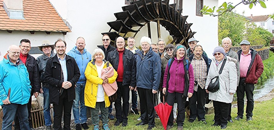 Eine Gruppe von Menschen steht vor einer alten Mühle mit großem Wasserrad. Die Menschen tragen warme Kleidung und Jacken. In der Mitte steht Bürgermeister Dr. Alexander Krey. Die Stimmung ist freundlich, alle lächeln. Im Hintergrund ist ein Bach und eine Brücke zu sehen. Der Himmel ist bewölkt.