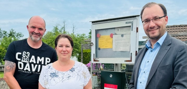 Drei Personen stehen draußen auf einem Weg. Sie schauen in die Kamera und lächeln. Rechts steht Bürgermeister Dr. Alexander Krey. Neben ihnen steht ein Spender für Hundekotbeutel. Der Spender ist schwarz und an einer Stange befestigt. Dahinter ist ein Schaukasten mit Zettel. Um den Weg wachsen viele Pflanzen. Der Himmel ist bewölkt.