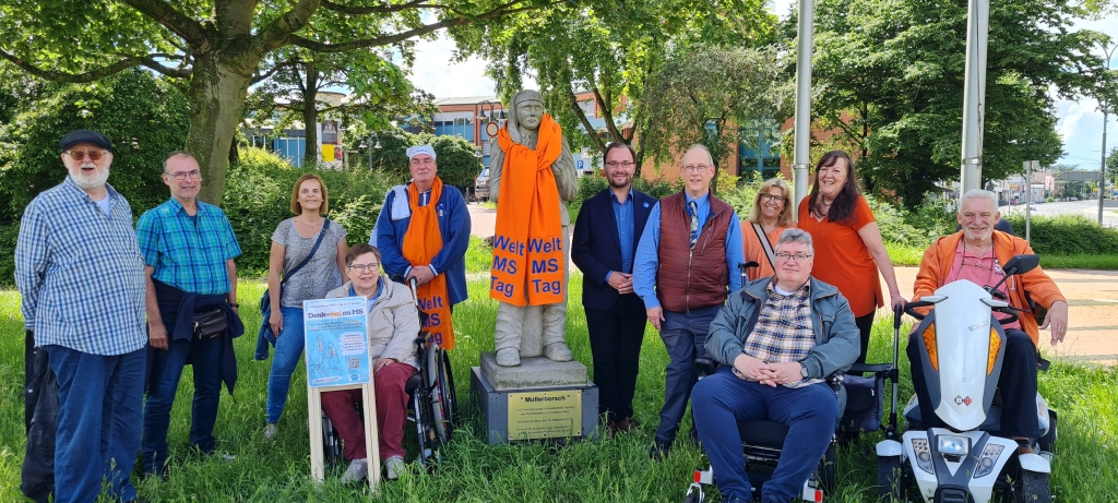 Eine Gruppe von Menschen steht auf einer Wiese. Viele tragen orangefarbene Schals mit der Aufschrift "Welt MS Tag". In der Mitte steht das Denkmal des Mühlheimer Müllerborsch mit einem orangefarbenen Schal. Manche Personen benutzen einen Rollstuhl. Im Hintergrund sind Bäume und ein Gebäude zu sehen.