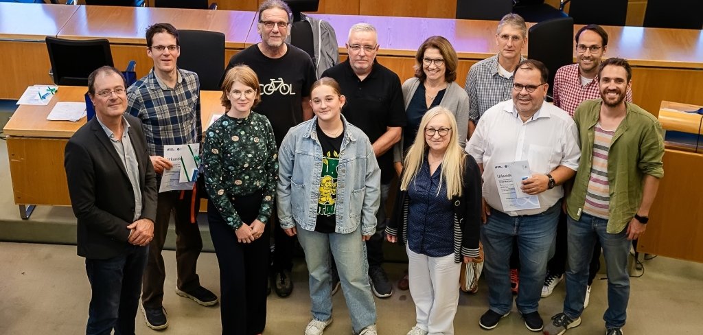 Das Bild zeigt eine Gruppe von Menschen in einem großen Raum. Der Raum hat viele Tische und Stühle. Die Menschen stehen zusammen und lächeln. Manche halten Urkunden in der Hand. Das Bild wurde bei der Ehrung für das Stadtradeln gemacht. Die Stadt Mühlheim hat die besten Fahrrad-Teams geehrt. Diese Teams sind besonders viel Fahrrad gefahren.